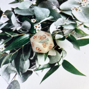 This double-poured D20 starts with a set of cream blanks with a hint of gold glitter throughout. Decorated with sprigs of a variety of plants, the second clear pour protects the images. Finished with gold inking.